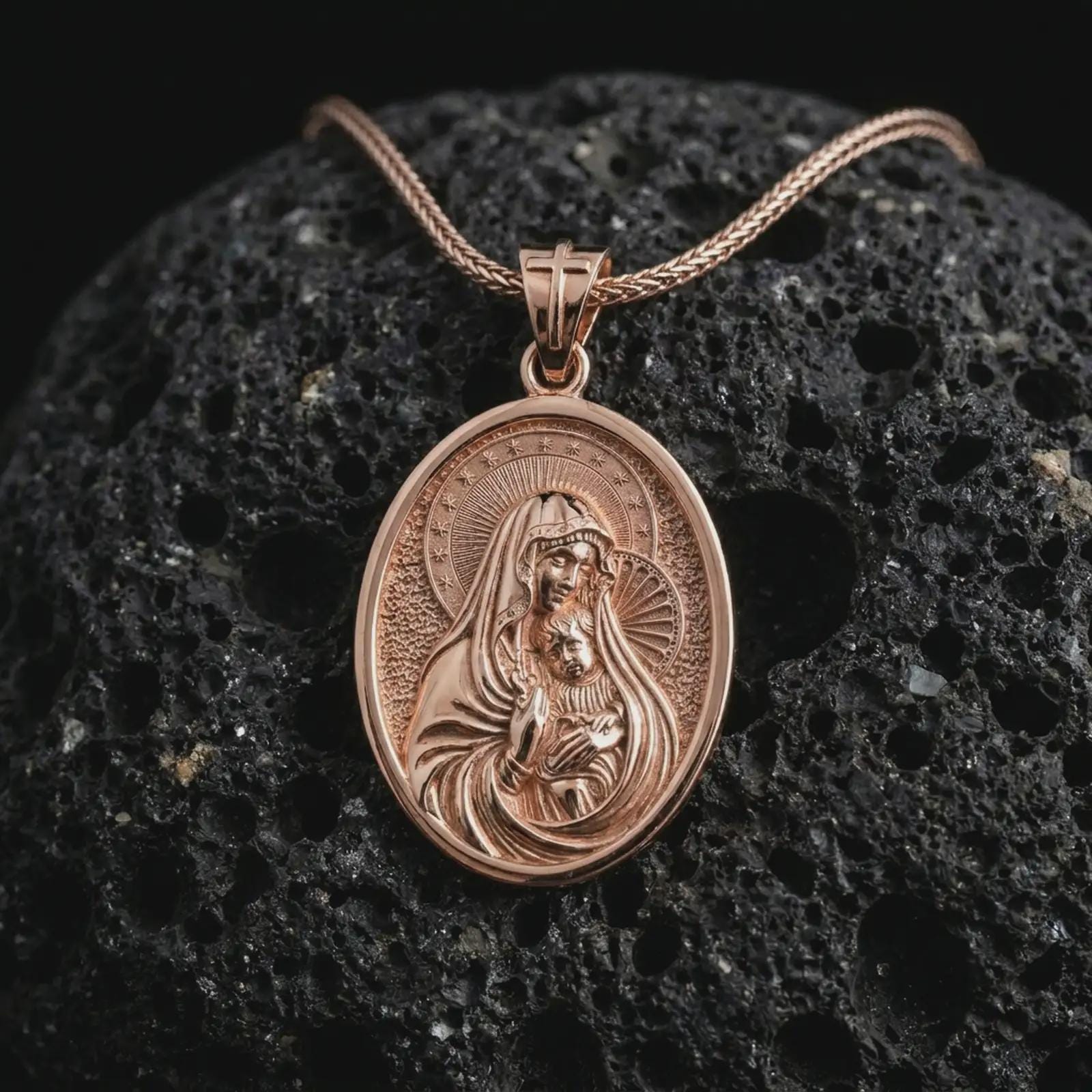 Rose gold plated Virgin Mary and baby Jesus pendant necklace on textured volcanic rock, detailed macro product shot with soft warm reflections and devotional mother-and-child symbolism.