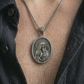 Load image into Gallery viewer, Oxidized Virgin Mary and baby Jesus pendant necklace worn on the chest, close-up lifestyle jewelry photo showing the religious medallion on a spiga chain with dark clothing.
