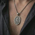 Load image into Gallery viewer, Oxidized Saint Anthony of Padua pendant necklace worn on the chest, close-up lifestyle jewelry photo showing the Catholic medallion on a spiga chain with dark clothing.
