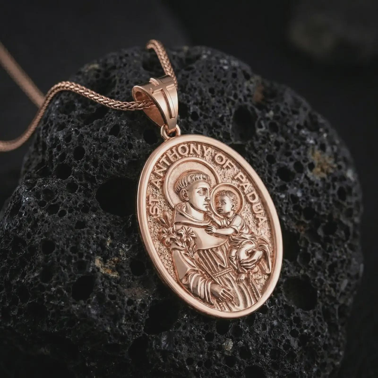 Rose gold plated Saint Anthony of Padua pendant necklace on textured volcanic rock, detailed macro product shot with soft warm reflections and devotional Saint Anthony design.