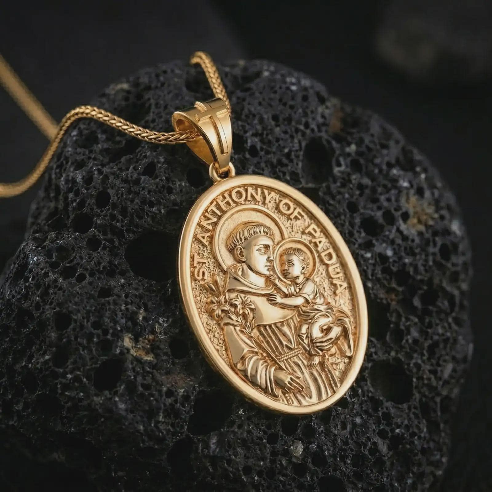 Gold plated Saint Anthony of Padua pendant necklace on textured volcanic rock, premium close-up studio composition emphasizing the saint holding baby Jesus and lily symbolism.