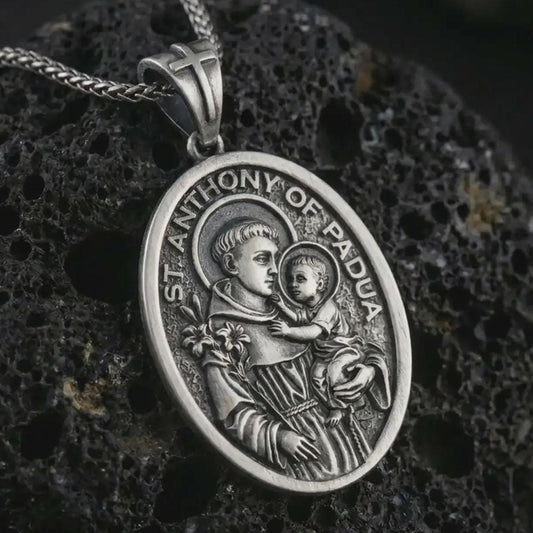 Oxidized Saint Anthony of Padua pendant necklace on textured volcanic rock, close-up studio shot highlighting the saint and child relief, lily detail, halo, and antique silver contrast.