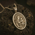 Load image into Gallery viewer, Bronze Saint Anthony of Padua pendant necklace on textured volcanic rock, close-up studio shot highlighting the saint and child relief, lily detail, halo, and antique silver contrast.
