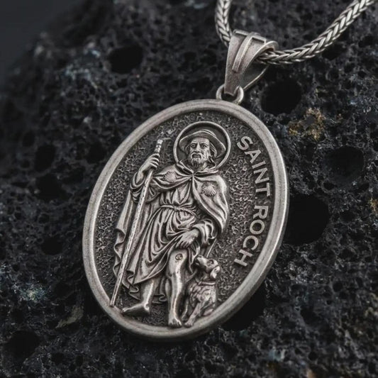 Oxidized Saint Roch pendant necklace on textured volcanic rock, close-up studio shot highlighting the saint relief, pilgrim staff, dog detail, and antique silver contrast.
