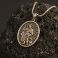 Load image into Gallery viewer, Bronze Saint Roch pendant necklace on textured volcanic rock, close-up studio shot highlighting the saint relief, pilgrim staff, dog detail, and antique silver contrast.
