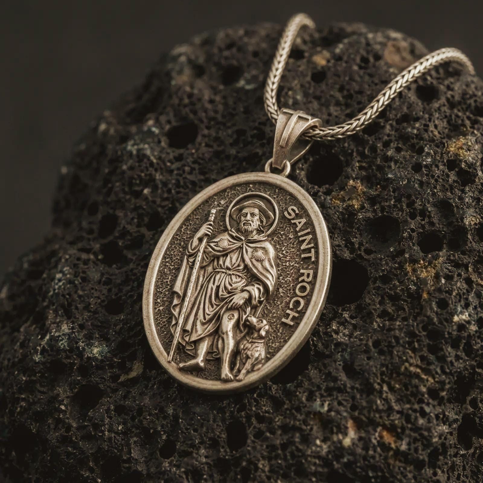 Bronze Saint Roch pendant necklace on textured volcanic rock, close-up studio shot highlighting the saint relief, pilgrim staff, dog detail, and antique silver contrast.