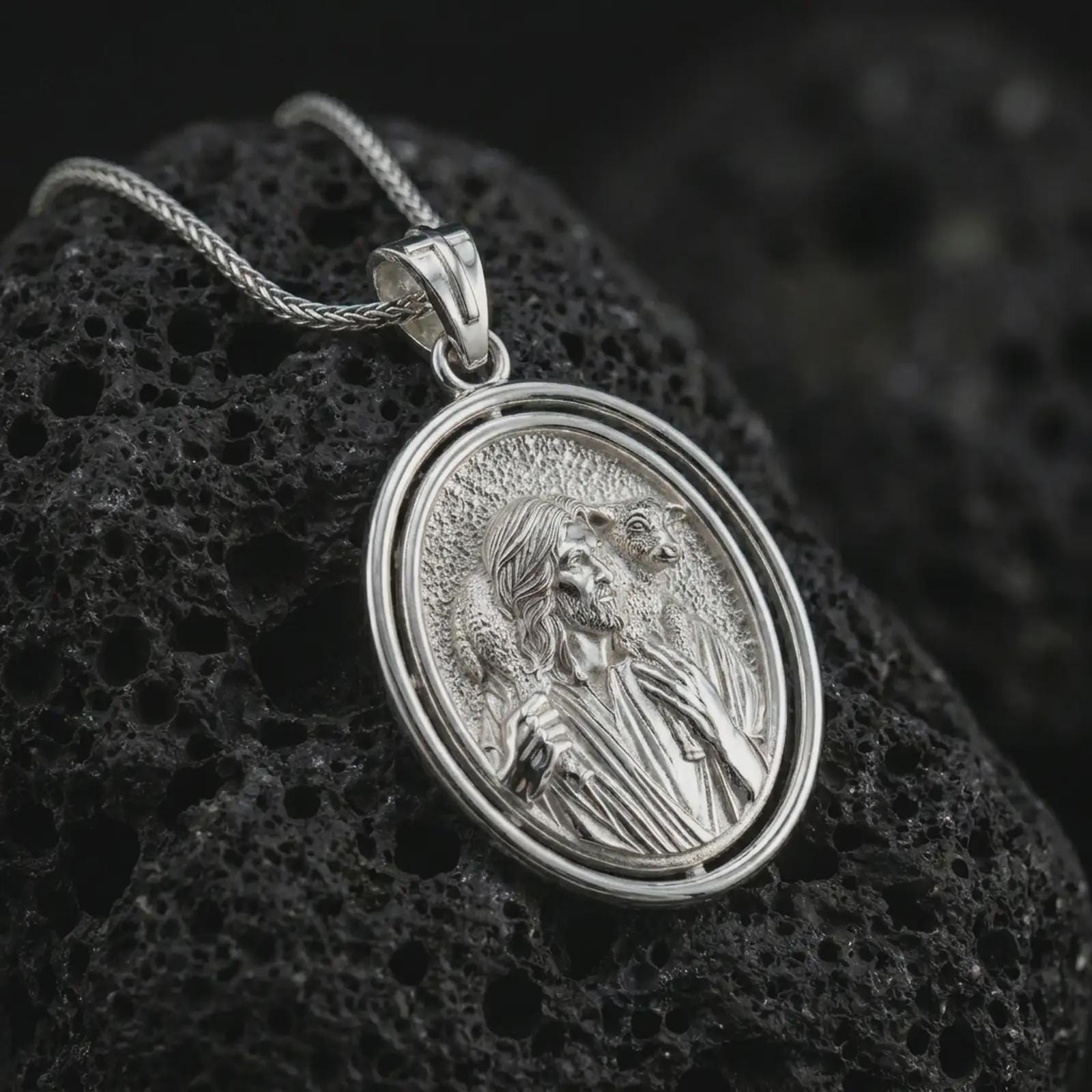 Polished silver spinning Good Shepherd pendant necklace on textured volcanic rock, centered macro jewelry photo with detailed shepherd relief and bright silver highlights.