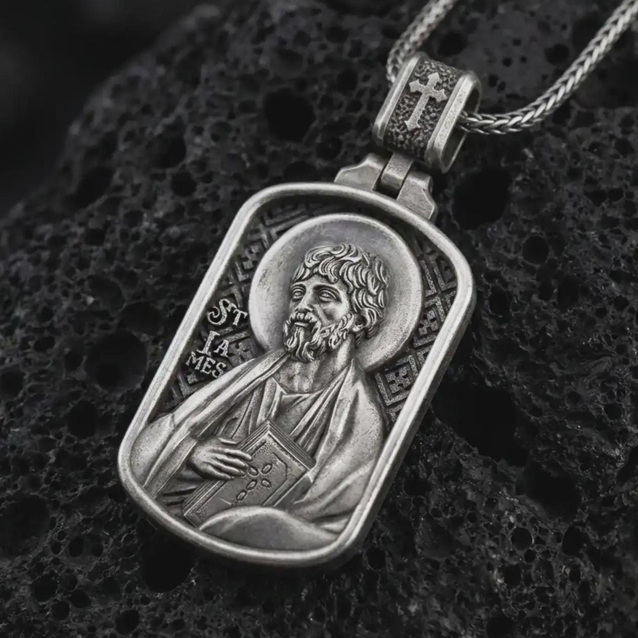 Oxidized Saint James the Apostle pendant necklace on textured volcanic rock, close-up studio shot highlighting the apostle relief, halo, book detail, and antique silver contrast.