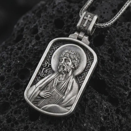 Oxidized Saint James the Apostle pendant necklace on textured volcanic rock, close-up studio shot highlighting the apostle relief, halo, book detail, and antique silver contrast.