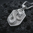 Load image into Gallery viewer, Polished silver cobra shield pendant necklace displayed on black volcanic rock, shown in a refined macro product shot with raised serpent body, detailed scales, and angular shield frame.
