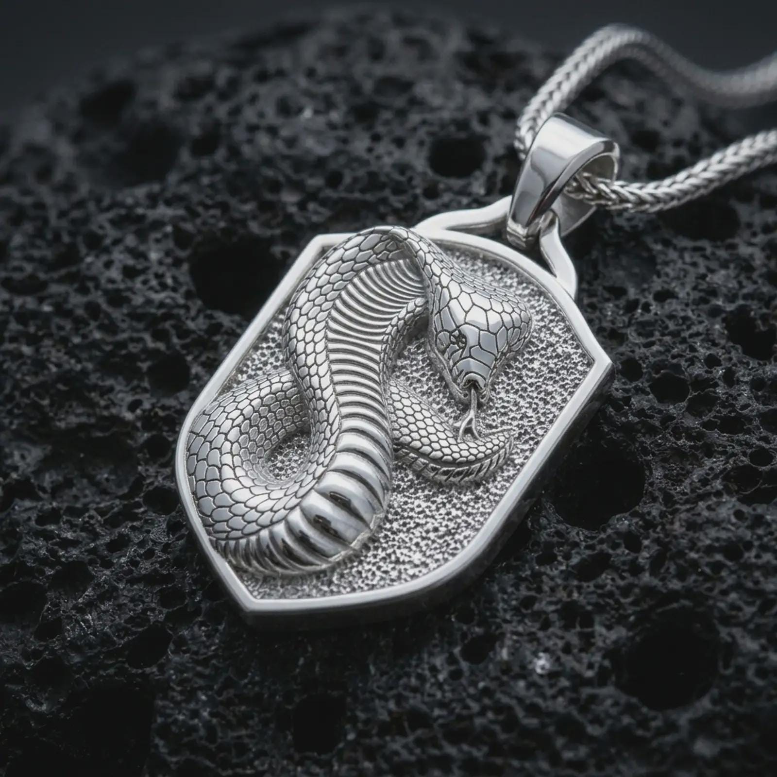 Polished silver cobra shield pendant necklace displayed on black volcanic rock, shown in a refined macro product shot with raised serpent body, detailed scales, and angular shield frame.