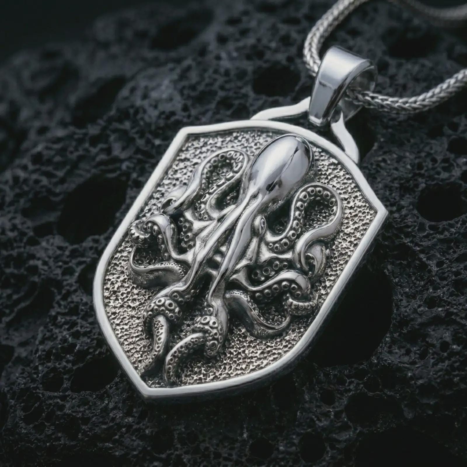 Polished silver kraken octopus shield pendant necklace displayed on black volcanic rock, shown in a refined macro product shot with curled tentacles, suction cup details, and angular shield frame.