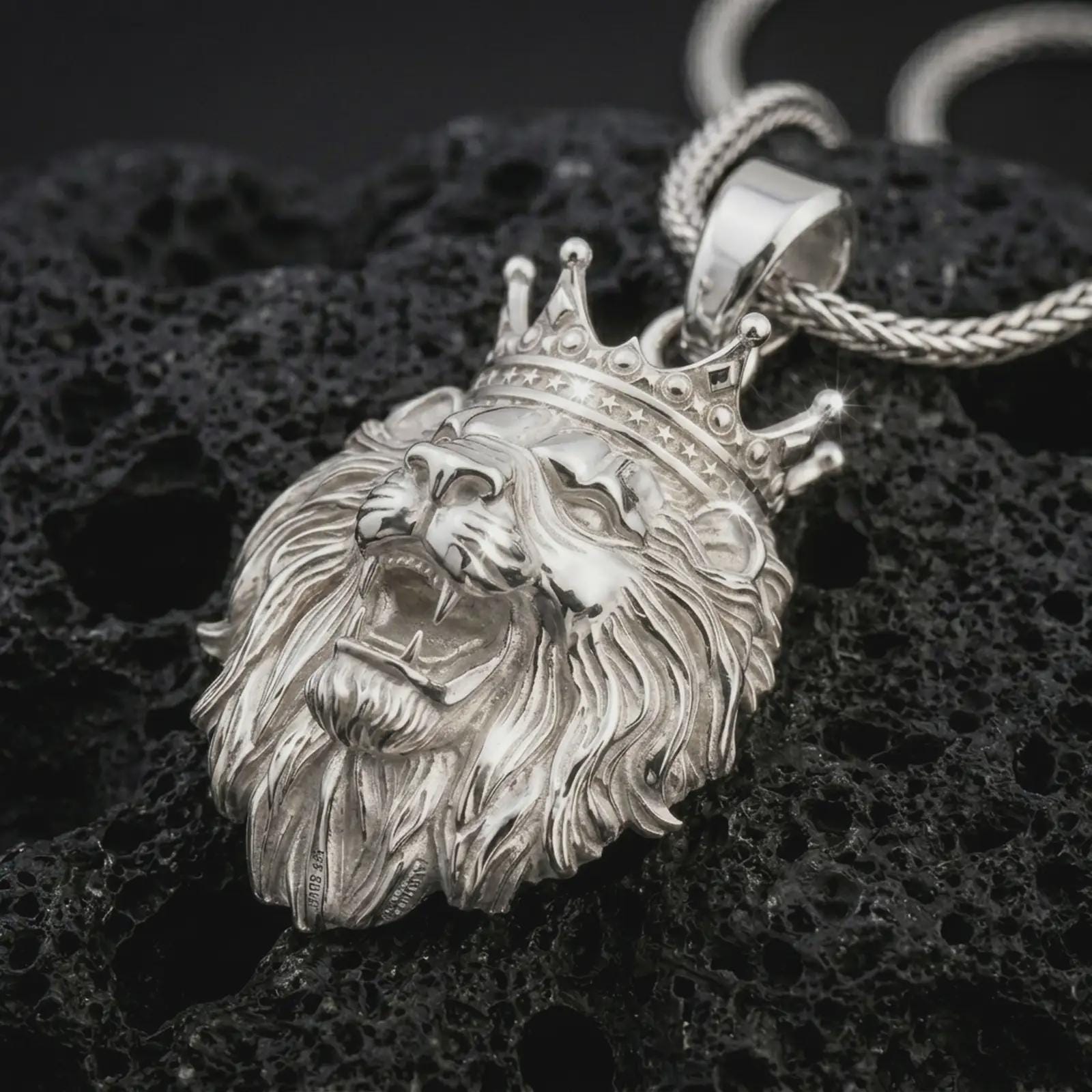 Polished silver crowned lion head pendant necklace displayed on black volcanic rock, shown in a refined macro product shot with royal crown detail, open roaring mouth, and flowing mane texture.