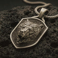 Load image into Gallery viewer, Bronze roaring bear shield pendant necklace with an oxidized finish resting on textured volcanic rock, captured in a close-up premium studio composition.
