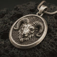Load image into Gallery viewer, Bronze ram head pendant necklace with an oxidized finish resting on textured volcanic rock, captured in a close-up premium studio composition.
