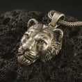 Load image into Gallery viewer, Bronze 3D lioness pendant necklace with an oxidized finish resting on textured volcanic rock, captured in a close-up premium studio composition.
