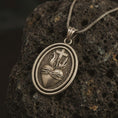 Load image into Gallery viewer, Bronze Sacred Heart pendant necklace on textured volcanic rock, close-up studio shot highlighting the flaming heart, crown of thorns, cross, and antique silver contrast.
