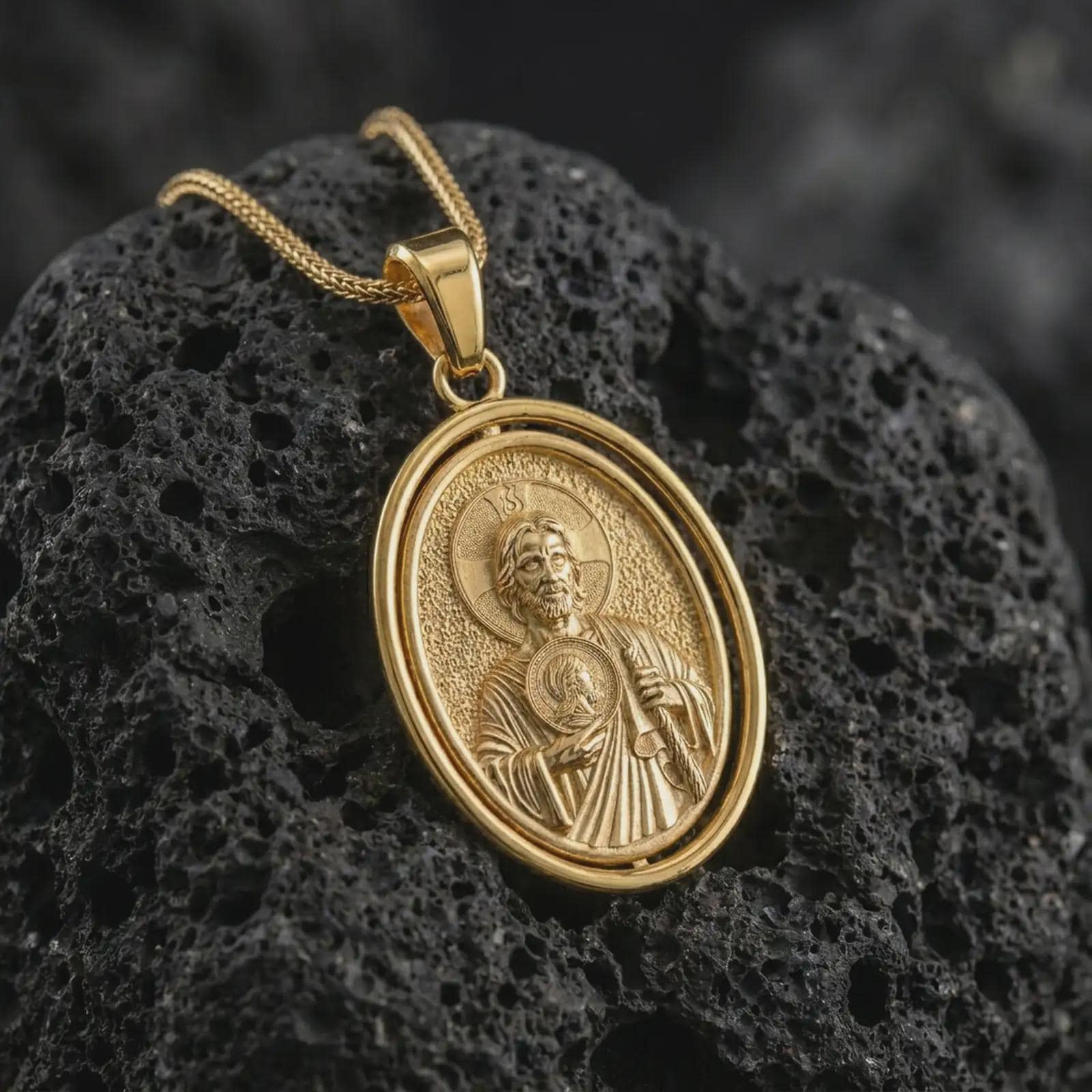 Gold plated Saint Jude pendant necklace on textured volcanic rock, premium close-up studio composition emphasizing the saint figure, halo, and sacred medallion symbolism.