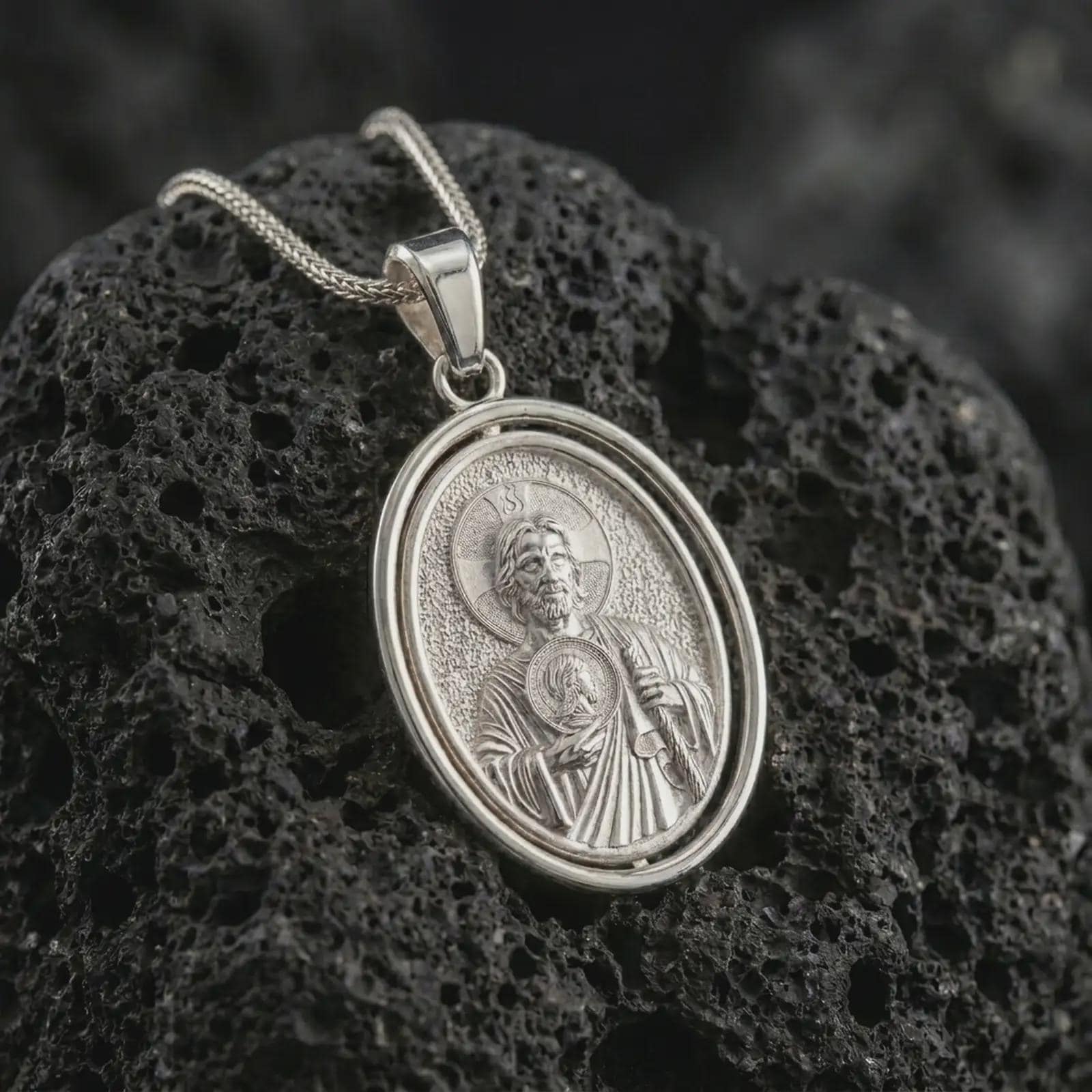 Polished silver Saint Jude pendant necklace on textured volcanic rock, centered macro jewelry photo with detailed Catholic medal engraving and bright silver highlights.