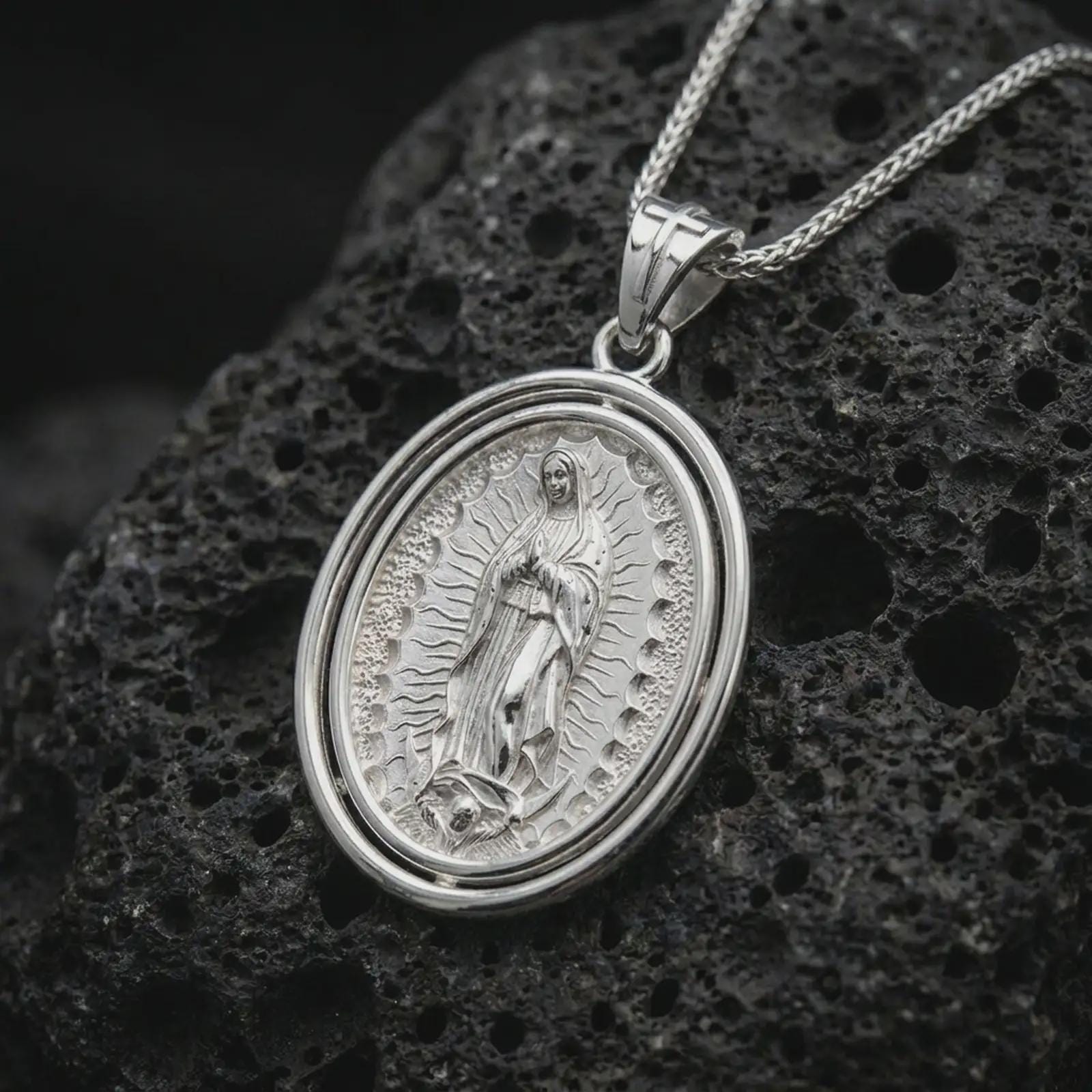 Polished silver Our Lady of Guadalupe spinner pendant necklace on textured volcanic rock, centered macro jewelry photo with detailed Catholic medal engraving and bright silver highlights.