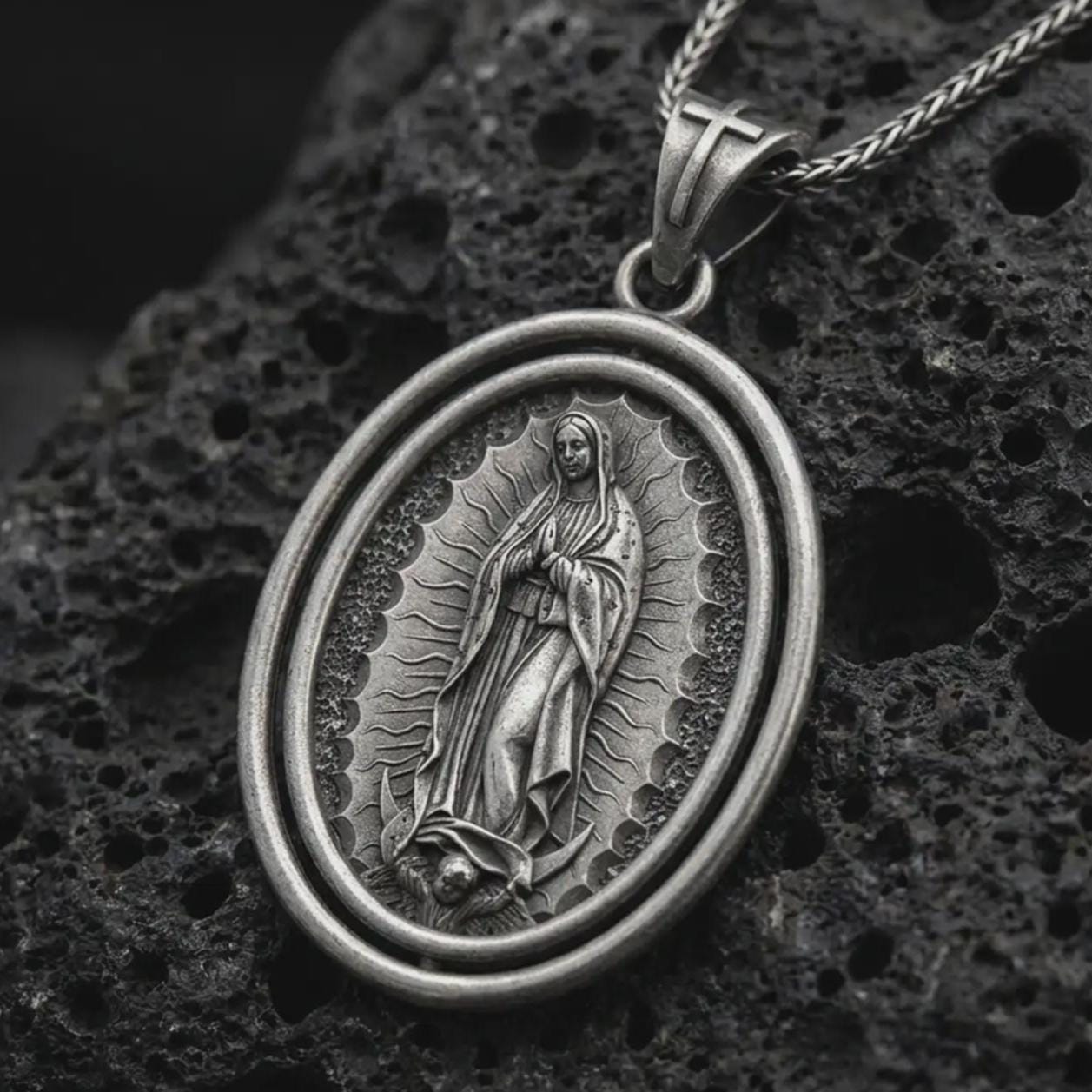 Oxidized Our Lady of Guadalupe spinner pendant necklace on textured volcanic rock, close-up studio shot highlighting the Virgin Mary relief, radiant halo, crescent moon, and antique silver contrast.