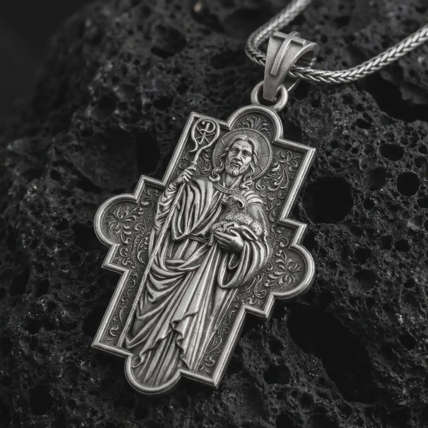 Oxidized Good Shepherd cross pendant necklace on textured volcanic rock, close-up studio shot highlighting the Jesus relief, lamb, shepherd staff, ornate cross frame, and antique silver contrast.