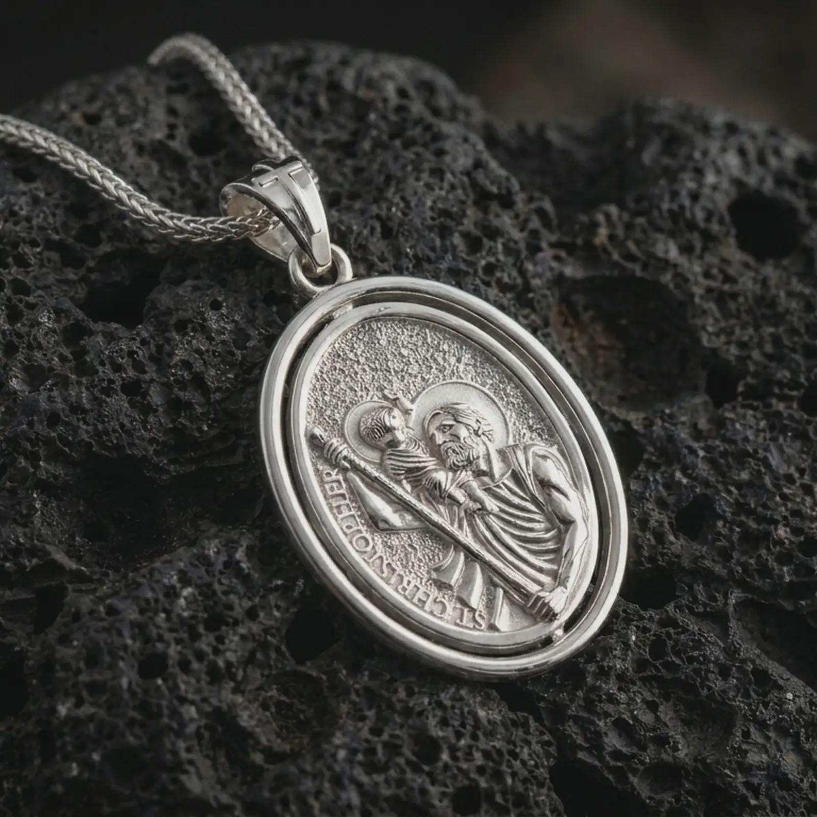 Polished silver Saint Christopher spinning pendant necklace on textured volcanic rock, centered macro jewelry photo with detailed Catholic medal relief and bright silver highlights.
