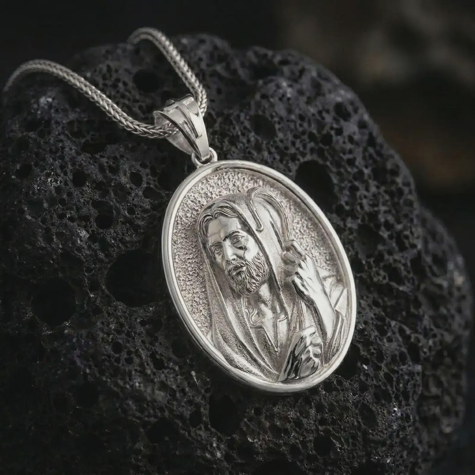 Polished silver Jesus Christ Good Shepherd pendant necklace on textured volcanic rock, centered macro jewelry photo with detailed Christian relief and bright silver highlights.