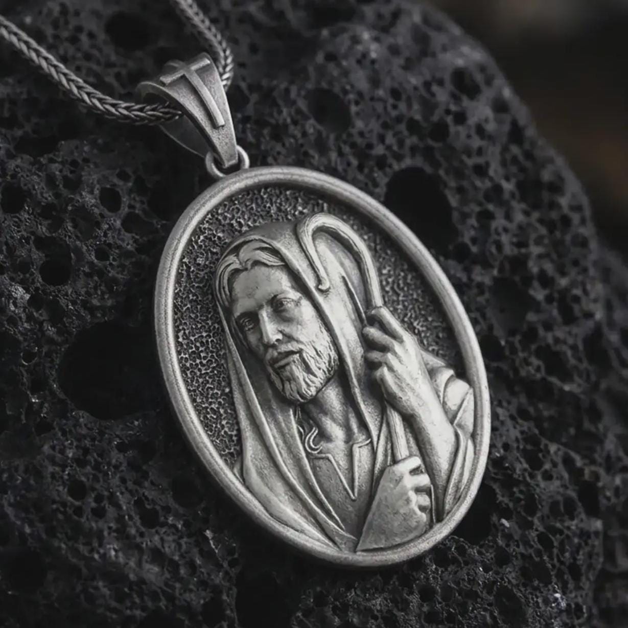 Oxidized Jesus Christ Good Shepherd pendant necklace on textured volcanic rock, close-up studio shot highlighting the shepherd portrait, hooded robe, staff detail, and antique silver contrast.
