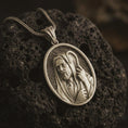Load image into Gallery viewer, Bronze Jesus Christ Good Shepherd pendant necklace on textured volcanic rock, close-up studio shot highlighting the shepherd portrait, hooded robe, staff detail, and antique silver contrast.
