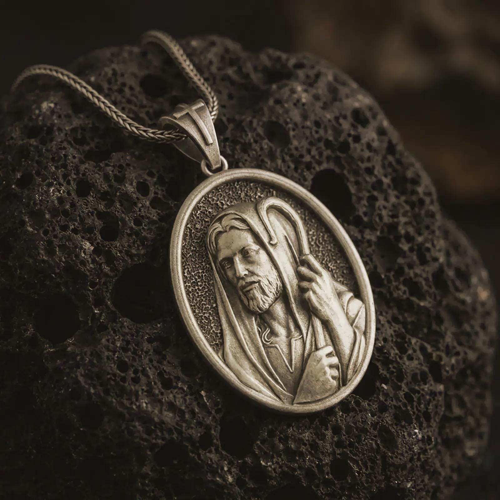 Bronze Jesus Christ Good Shepherd pendant necklace on textured volcanic rock, close-up studio shot highlighting the shepherd portrait, hooded robe, staff detail, and antique silver contrast.