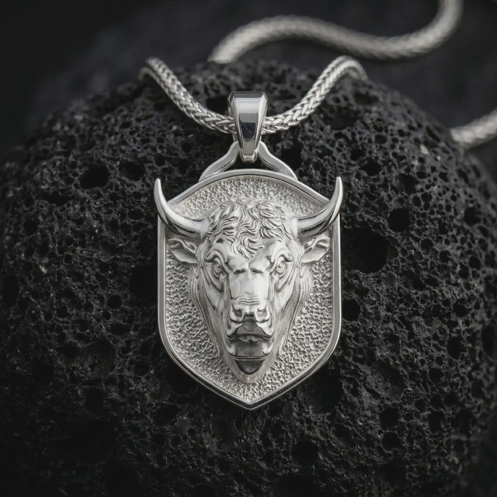 Polished silver bull head shield pendant necklace displayed on black volcanic rock, shown in a refined macro product shot with sharp horns, intense facial sculpting, and geometric shield frame.