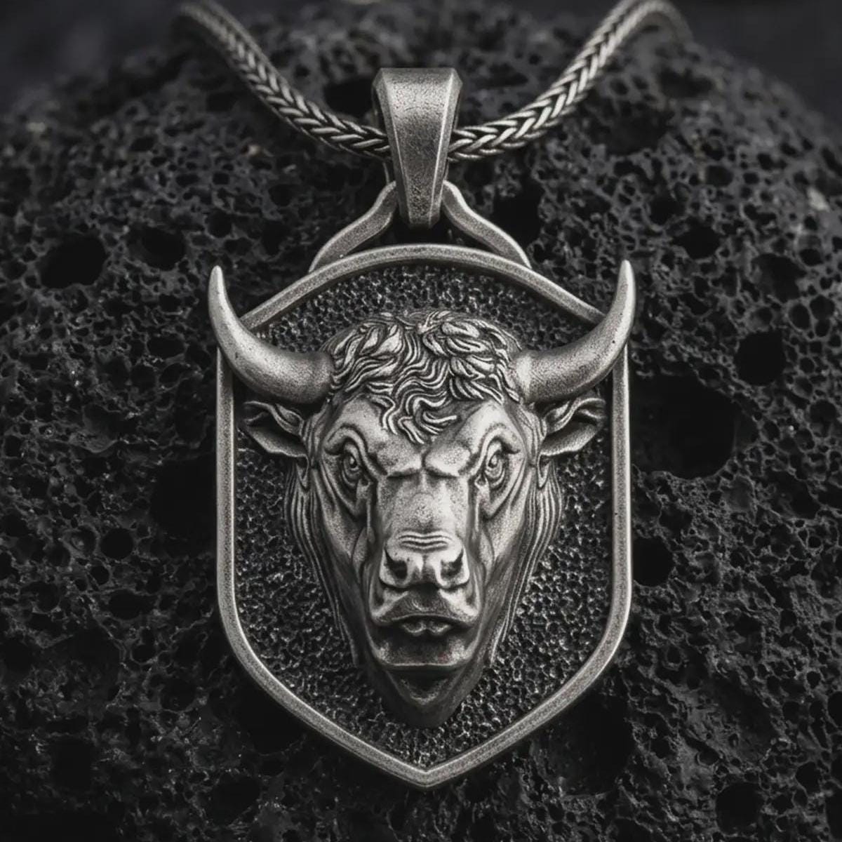 Sterling silver bull head shield pendant necklace with an oxidized finish resting on textured volcanic rock, captured in a close-up premium studio composition.