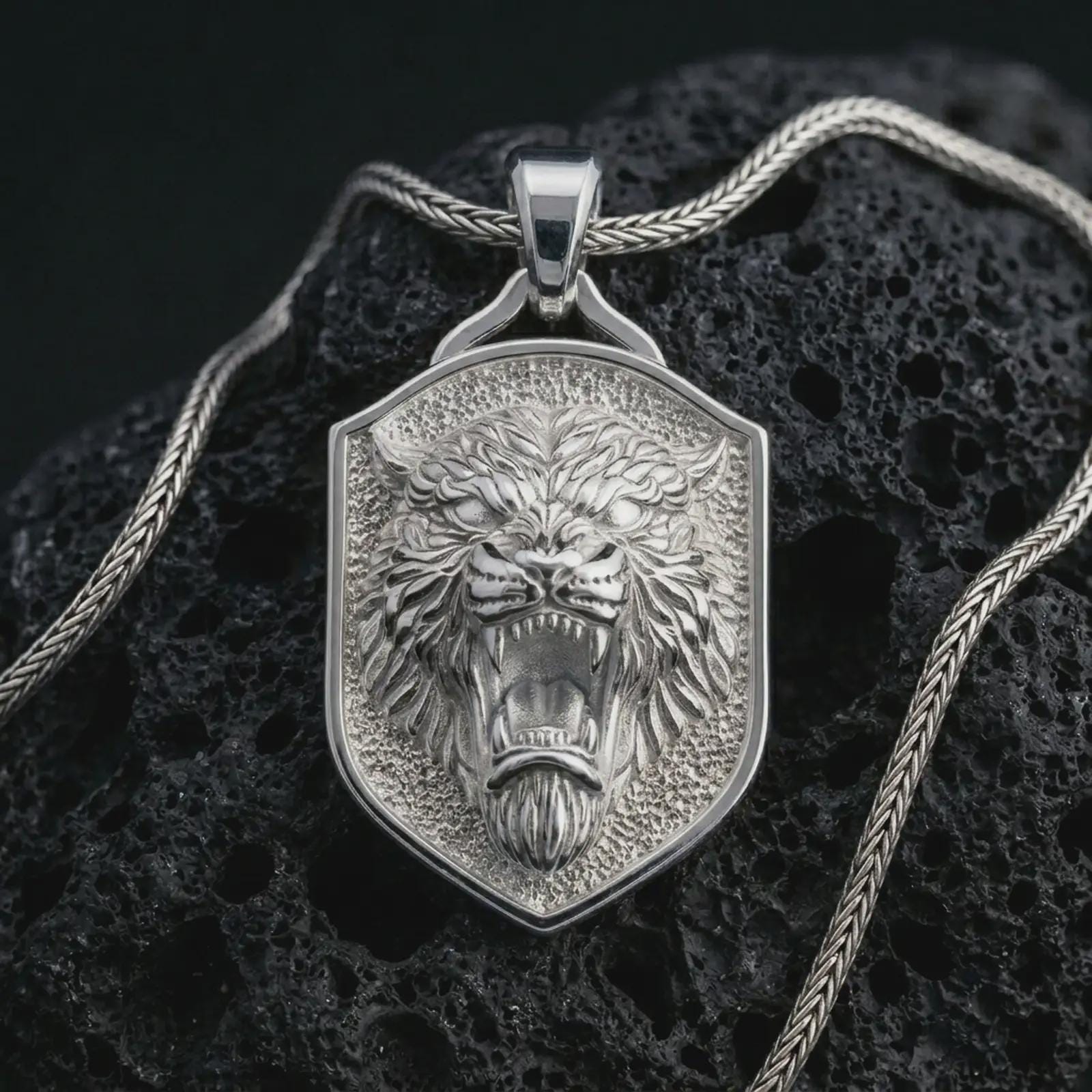 Polished silver roaring lion shield pendant necklace displayed on black volcanic rock, shown in a refined macro product shot with fierce open jaws, fang detail, and sculpted shield frame.
