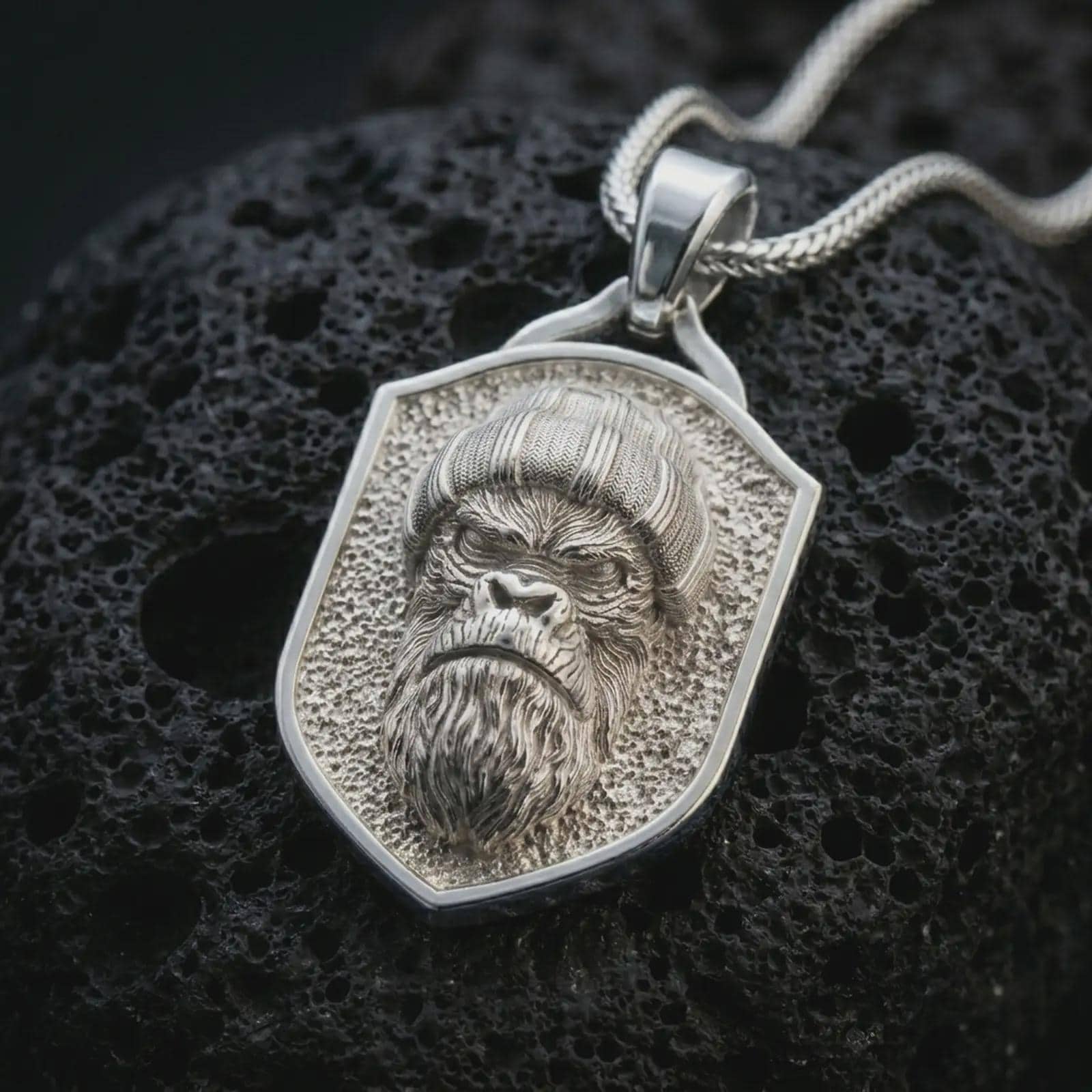 Polished silver gorilla shield pendant necklace displayed on black volcanic rock, shown in a refined macro product shot with stern ape face relief, knit beanie detail, and angular shield frame.