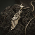 Load image into Gallery viewer, Bronze raven pendant necklace with an oxidized finish resting on textured volcanic rock, captured in a close-up premium studio composition.
