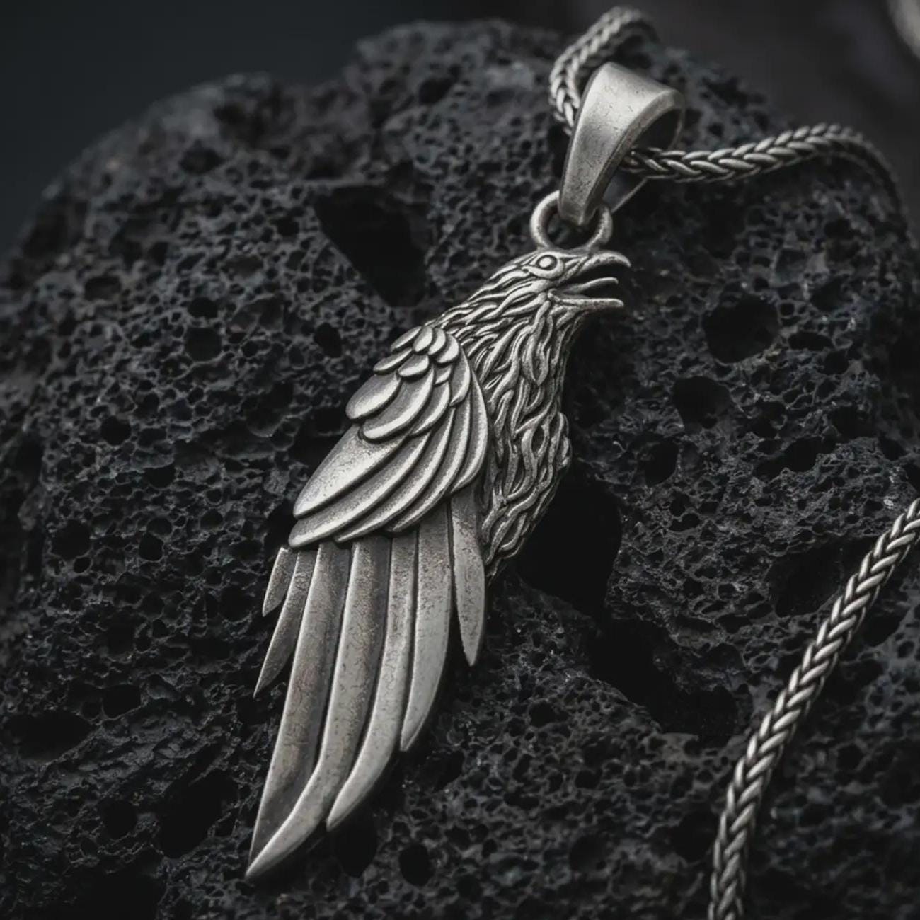 Sterling silver raven pendant necklace with an oxidized finish resting on textured volcanic rock, captured in a close-up premium studio composition.