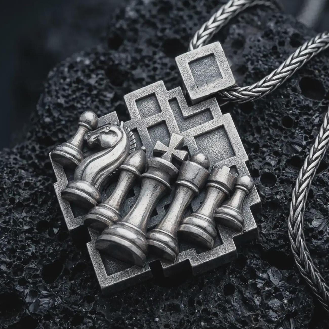 Sterling silver chessboard pendant necklace with an oxidized finish resting on textured volcanic rock, captured in a close-up premium studio composition.