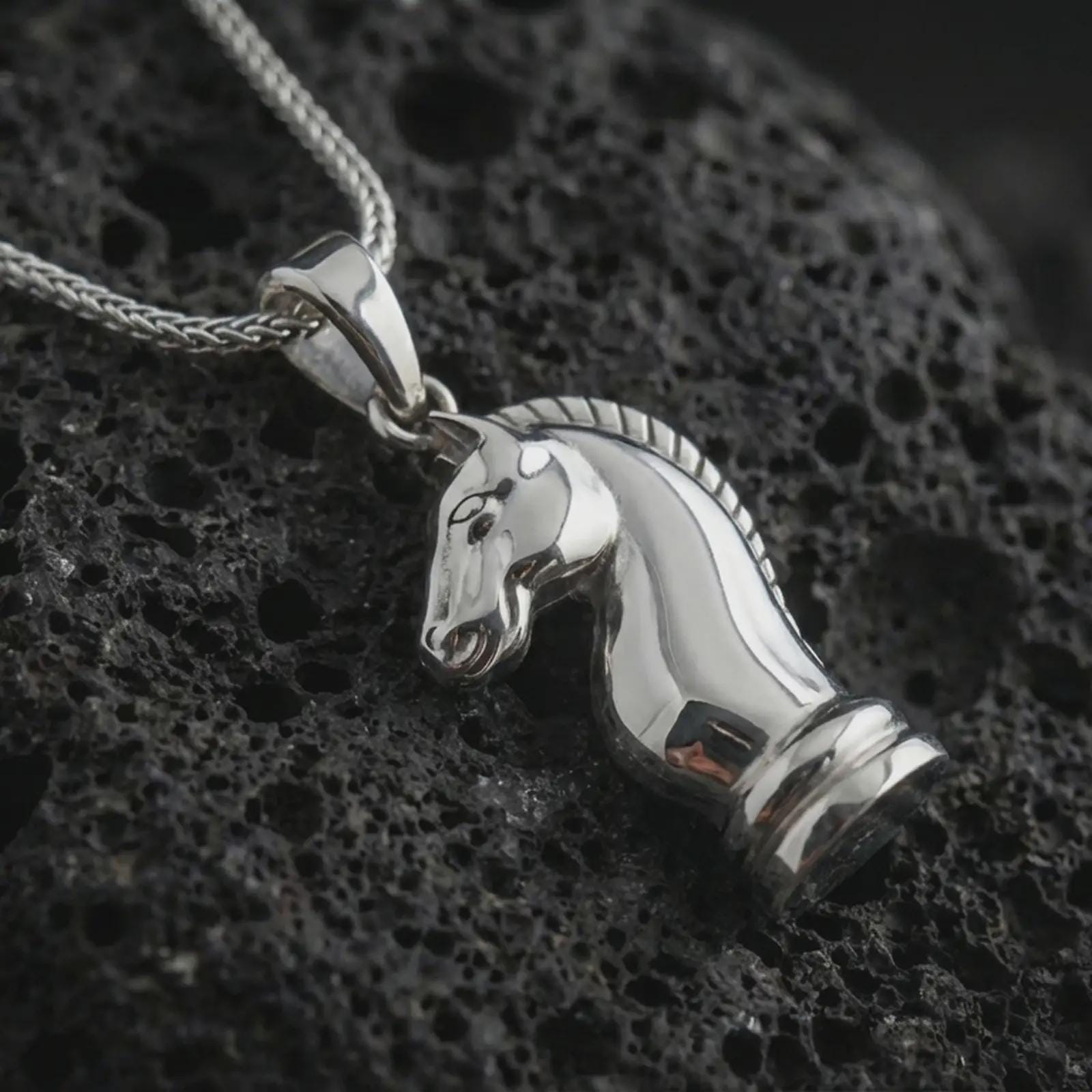 Polished silver knight chess piece pendant necklace displayed on black volcanic rock, shown in a refined macro product shot with sculpted horse head profile, smooth contours, and minimalist game-inspired design.