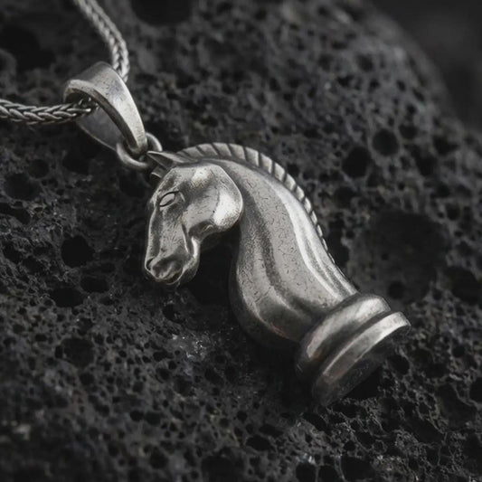 Sterling silver knight chess piece pendant necklace with an oxidized finish resting on textured volcanic rock, captured in a close-up premium studio composition.