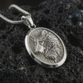 Load image into Gallery viewer, Polished silver wolf medallion pendant necklace displayed on black volcanic rock, shown in a refined macro product shot with side-profile wolf relief, flowing fur detail, and oval frame.
