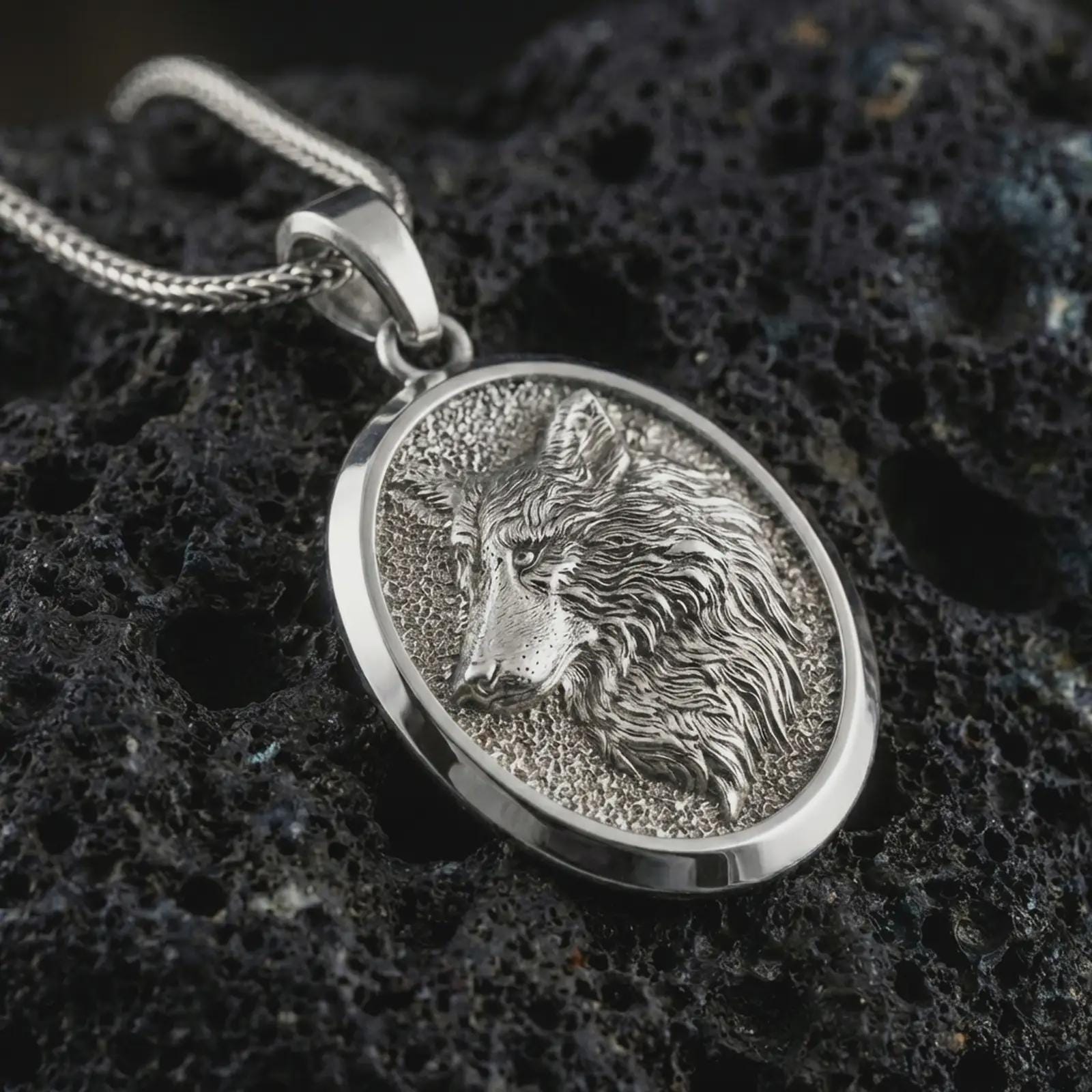 Polished silver wolf medallion pendant necklace displayed on black volcanic rock, shown in a refined macro product shot with side-profile wolf relief, flowing fur detail, and oval frame.