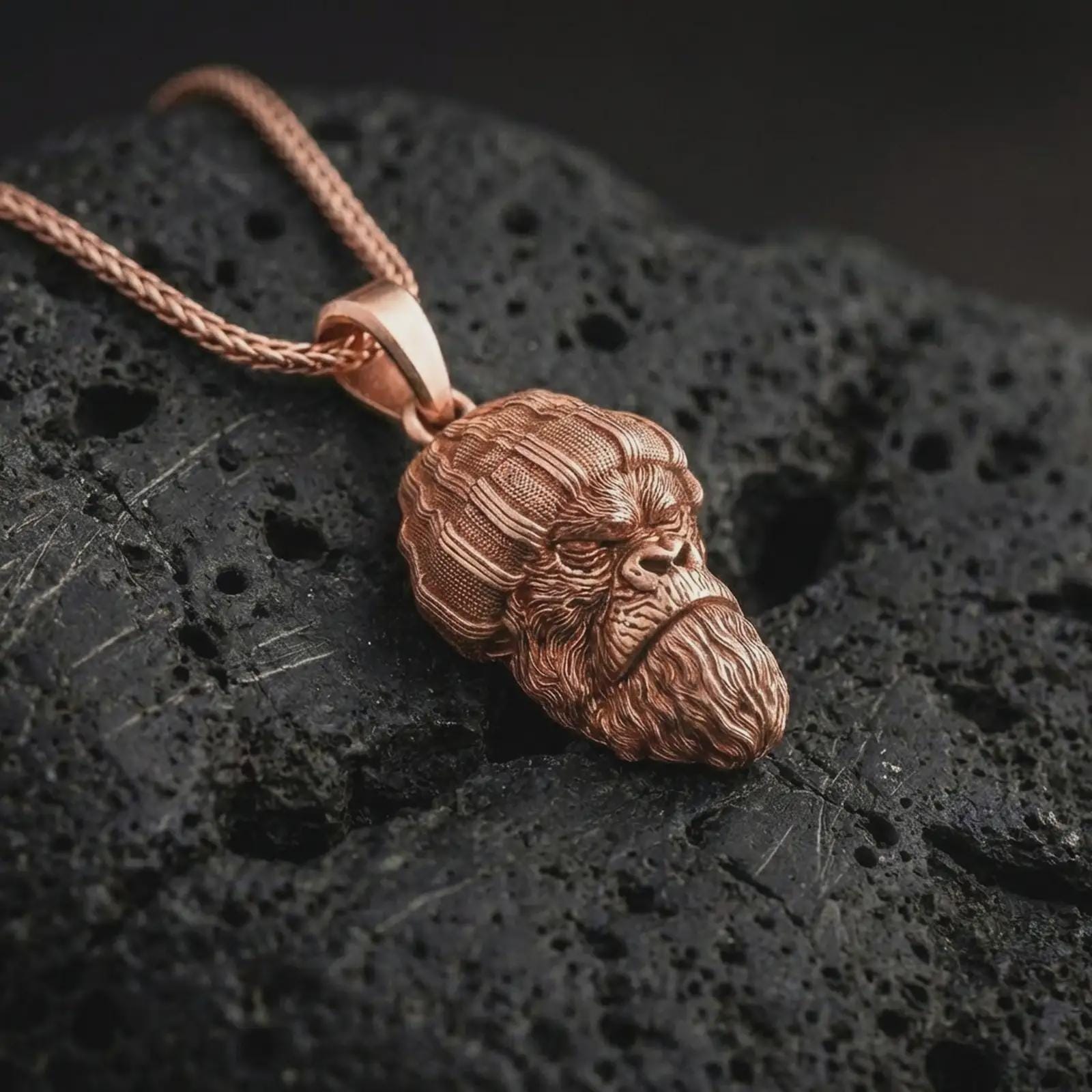 Rose gold plated gorilla head pendant necklace on dark volcanic rock, photographed in a detailed premium studio macro view.
