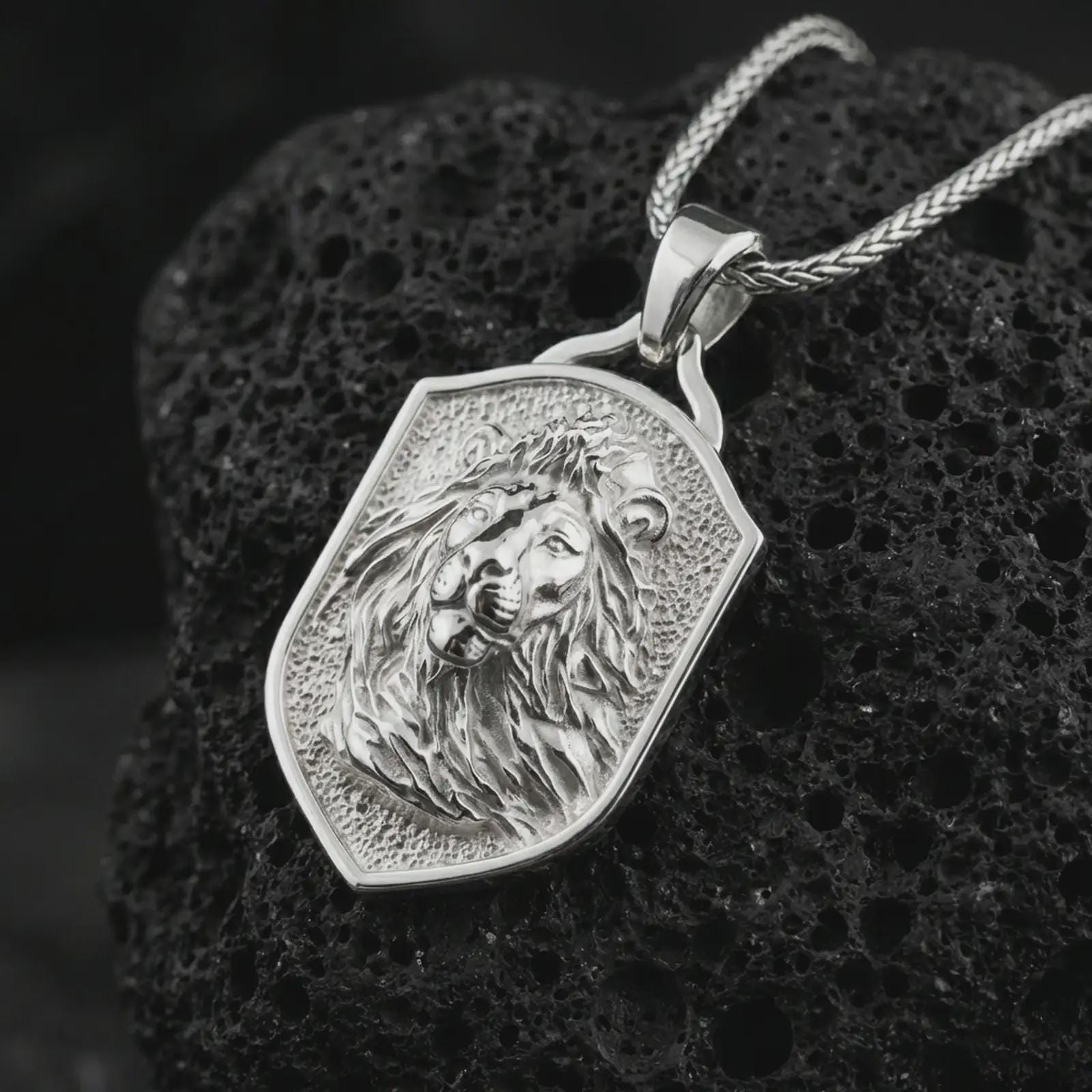 Polished silver lion shield pendant necklace displayed on black volcanic rock, shown in a refined macro product shot with windswept mane detail, regal facial sculpting, and angular shield frame.