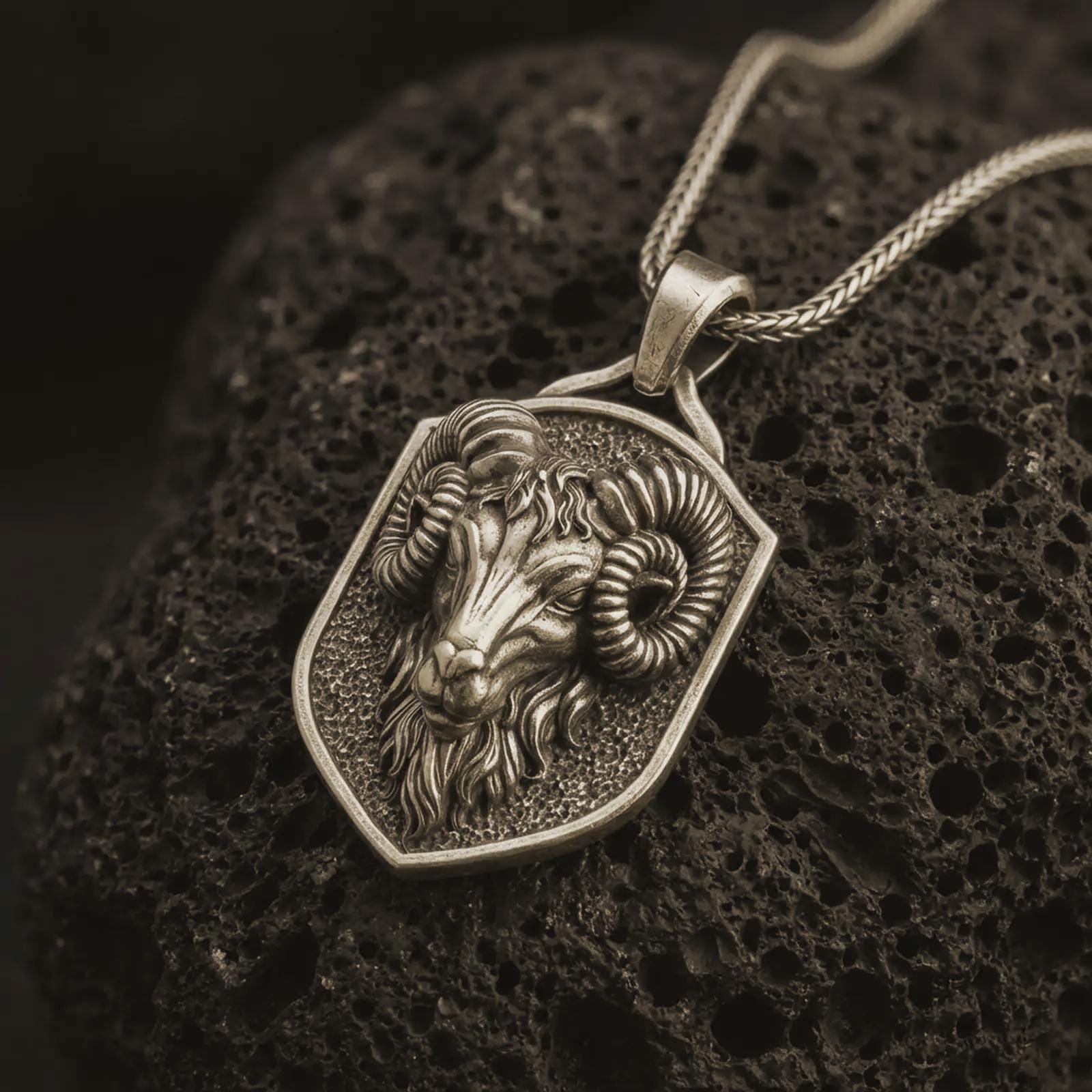 Bronze ram head shield pendant necklace with an oxidized finish resting on textured volcanic rock, captured in a close-up premium studio composition.