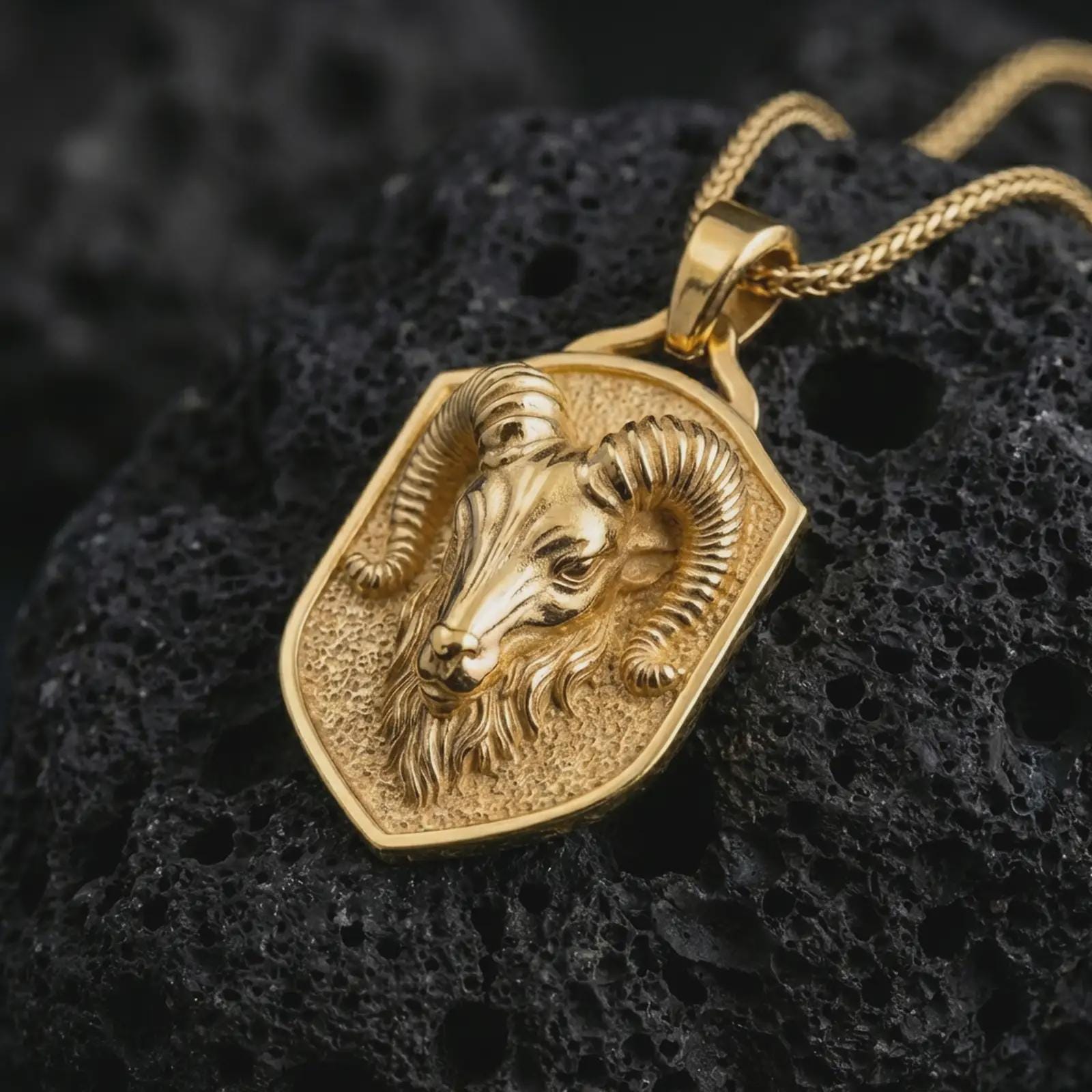Ram head shield pendant necklace with a gold plated finish displayed on porous volcanic stone, photographed in a refined macro composition with bold animal symbolism and shield contours.