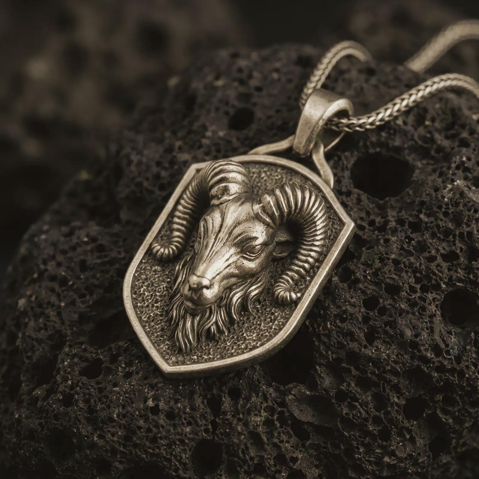 Ram head shield pendant necklace in oxidized Bronze resting on black volcanic stone, shown in a detailed macro product photograph with curved horns and a textured shield frame.
