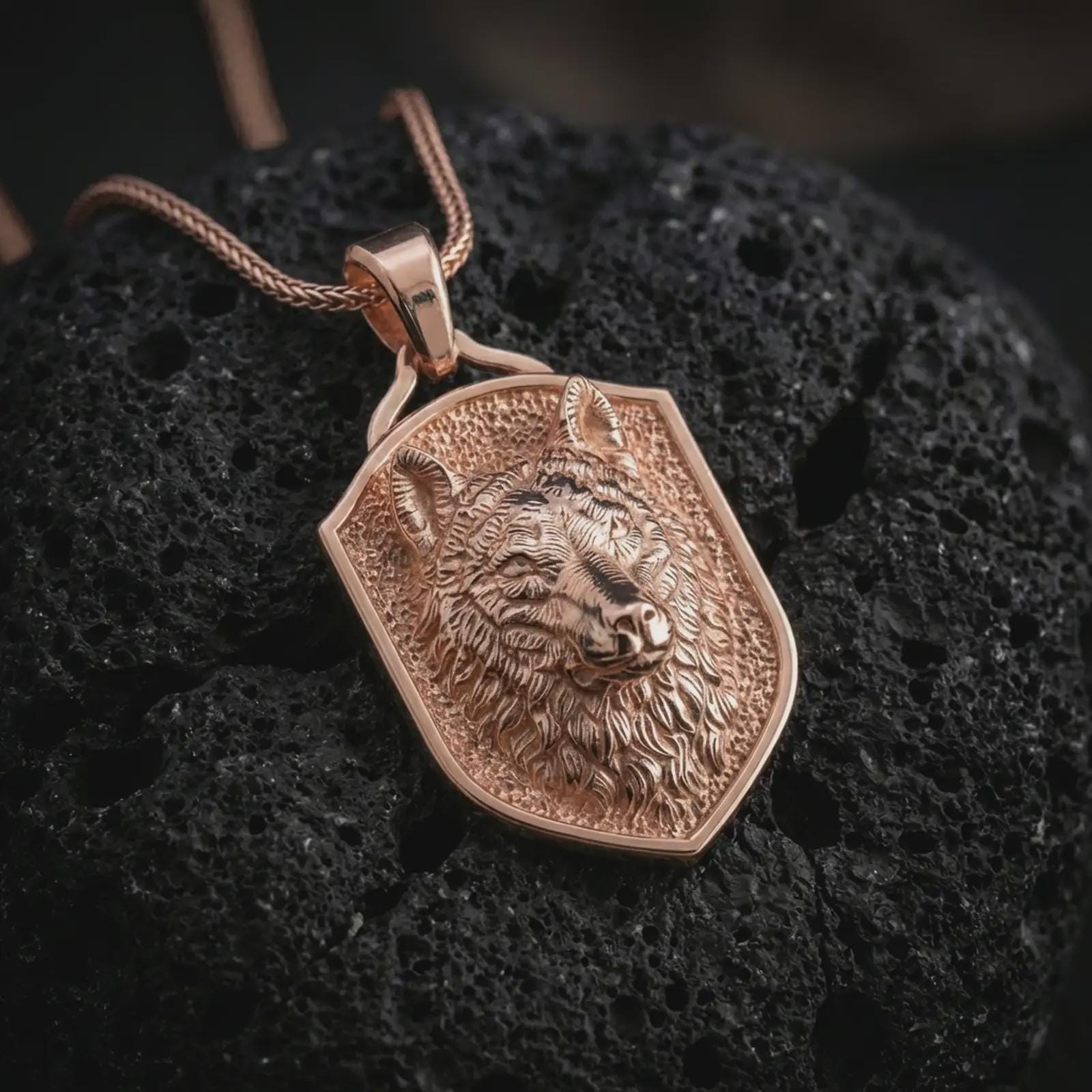 Wolf shield pendant necklace with a rose gold plated finish resting on textured black volcanic rock, shown in a premium close-up image with rich fur texture and a sharp shield outline.