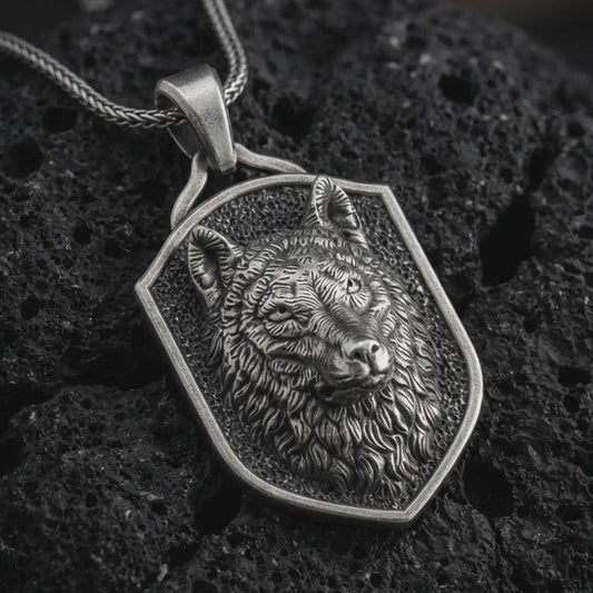 Wolf shield pendant necklace in oxidized sterling silver resting on black volcanic stone, shown in a detailed macro product photograph with textured fur, alert ears, and an angular shield frame.