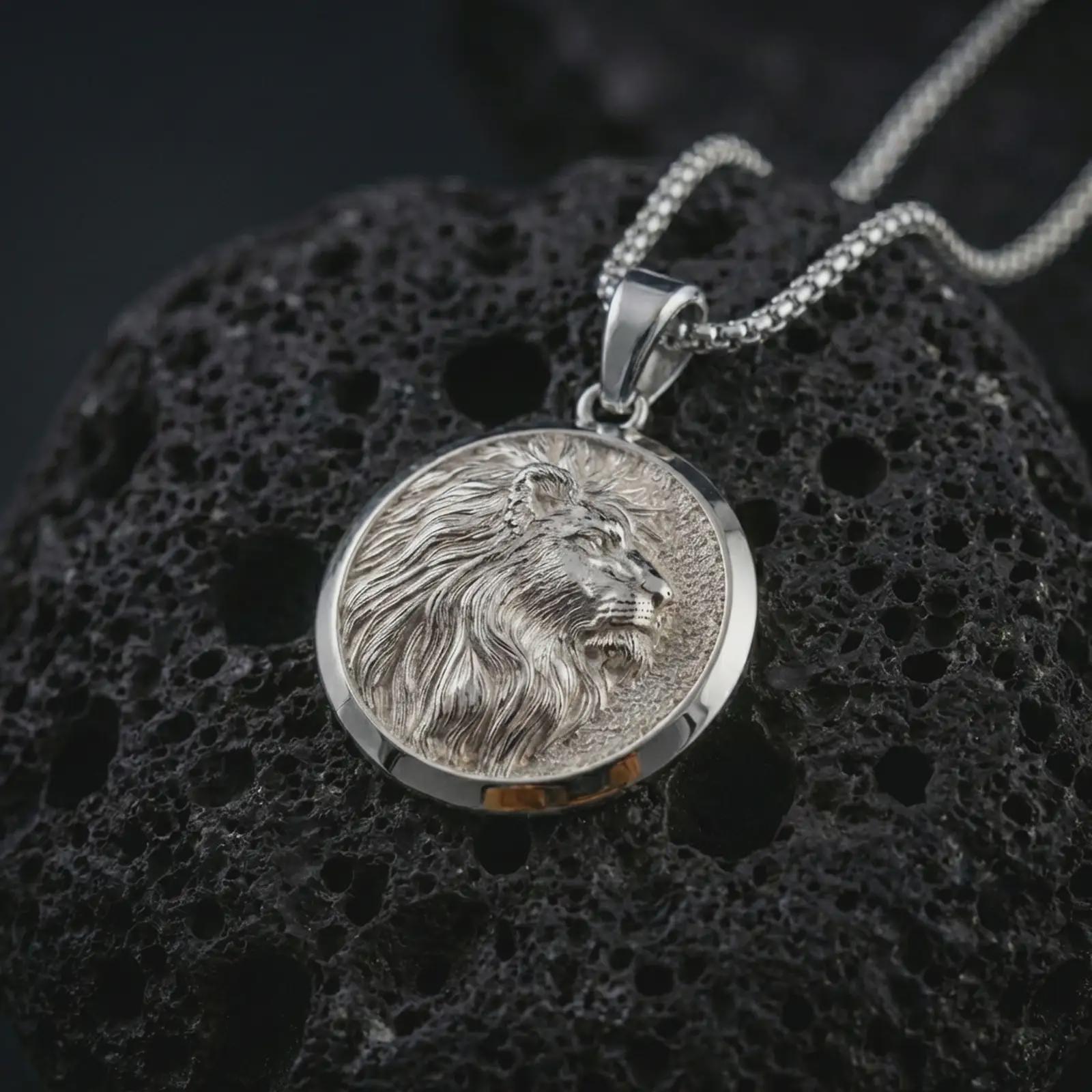Polished silver lion medallion pendant necklace displayed on black volcanic rock, shown in a refined macro product shot with side profile lion relief, flowing mane detail, and round frame.